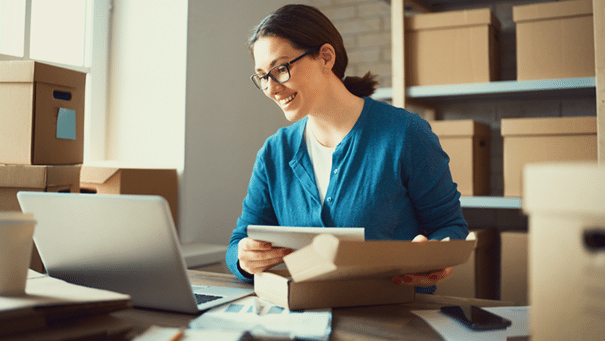 Femme dans un entrepôt qui consulte son ordinateur tout en préparant un colis.